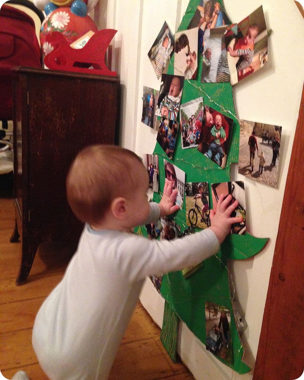 A toddler in pajamas reaches for a wall-mounted Christmas tree made of photos, set against a wooden floor and white wall.