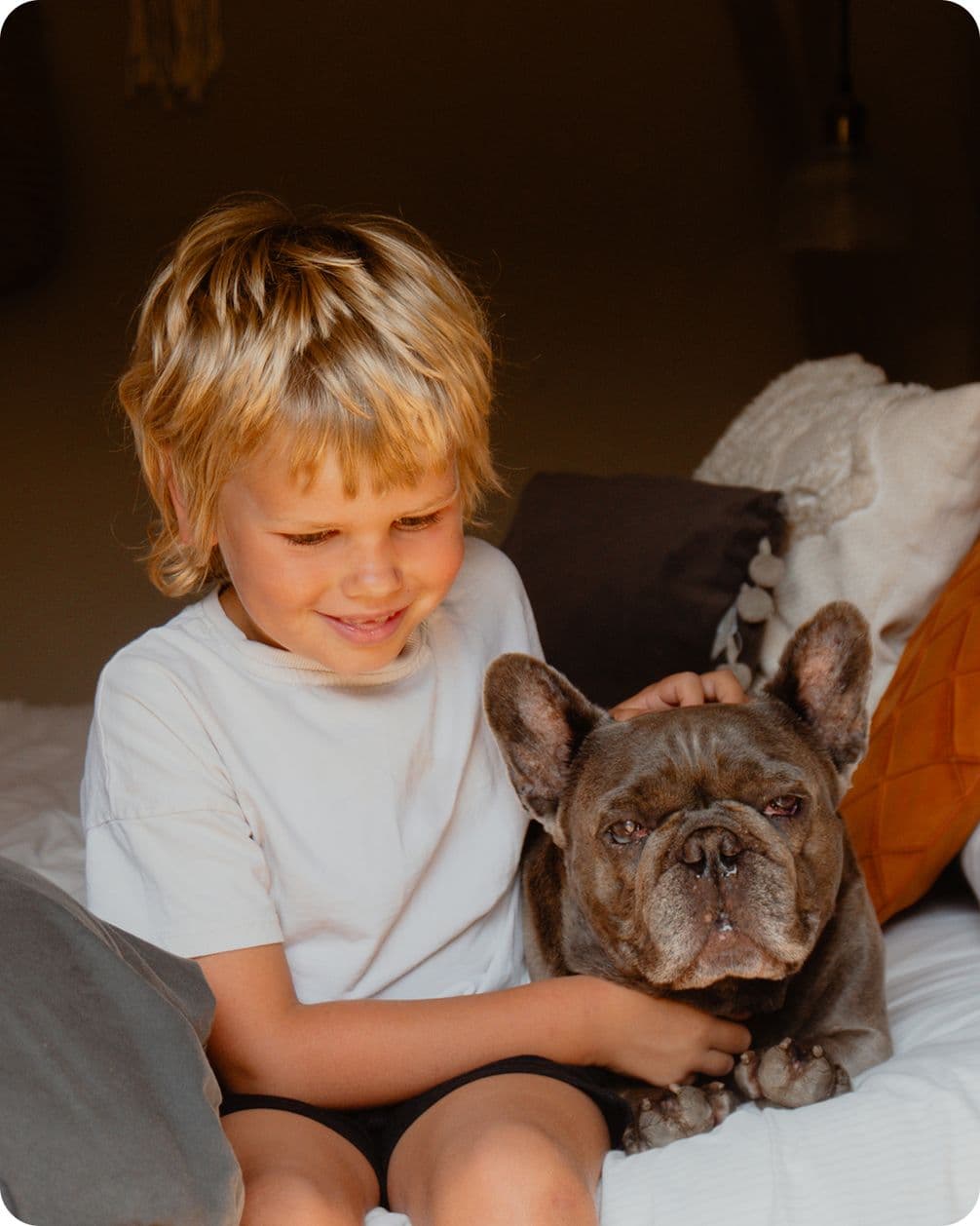 Young boy with blond hair sits on a bed, smiling and petting a relaxed French Bulldog.