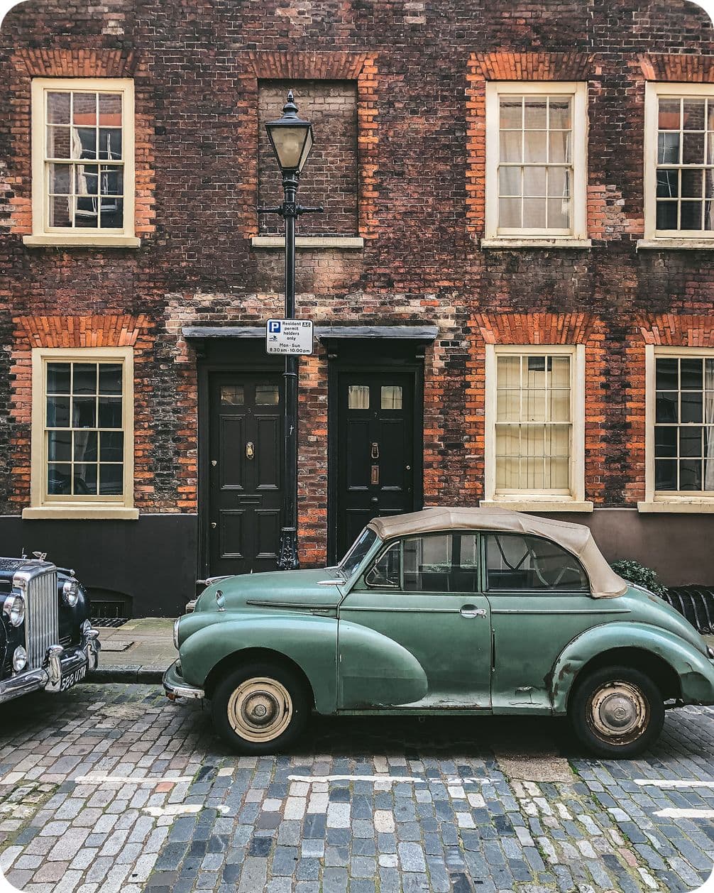 Vintage green car parked on a cobblestone street in front of an old brick building with a classic streetlamp and adjacent black doors.