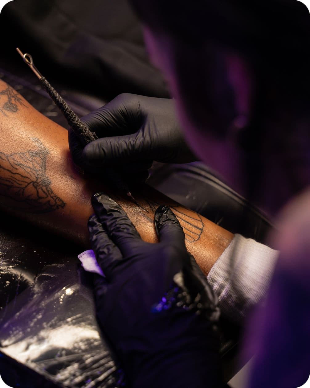 Tattoo artist wearing black gloves, focused on inking a detailed design on a person's forearm, under soft lighting.
