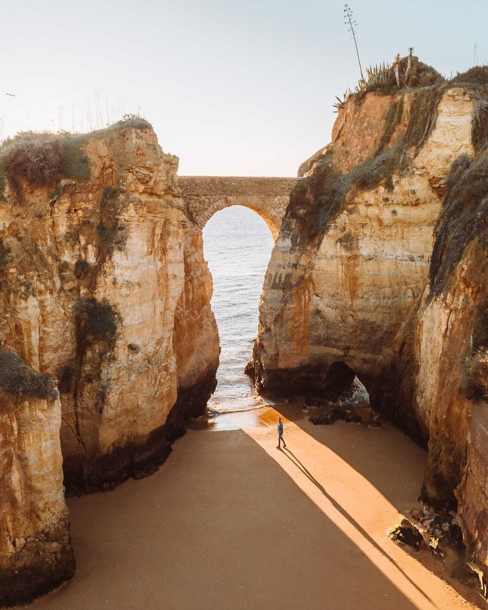A person stands on a sunlit beach beneath a stone archway between two cliffs, with the ocean visible in the background.