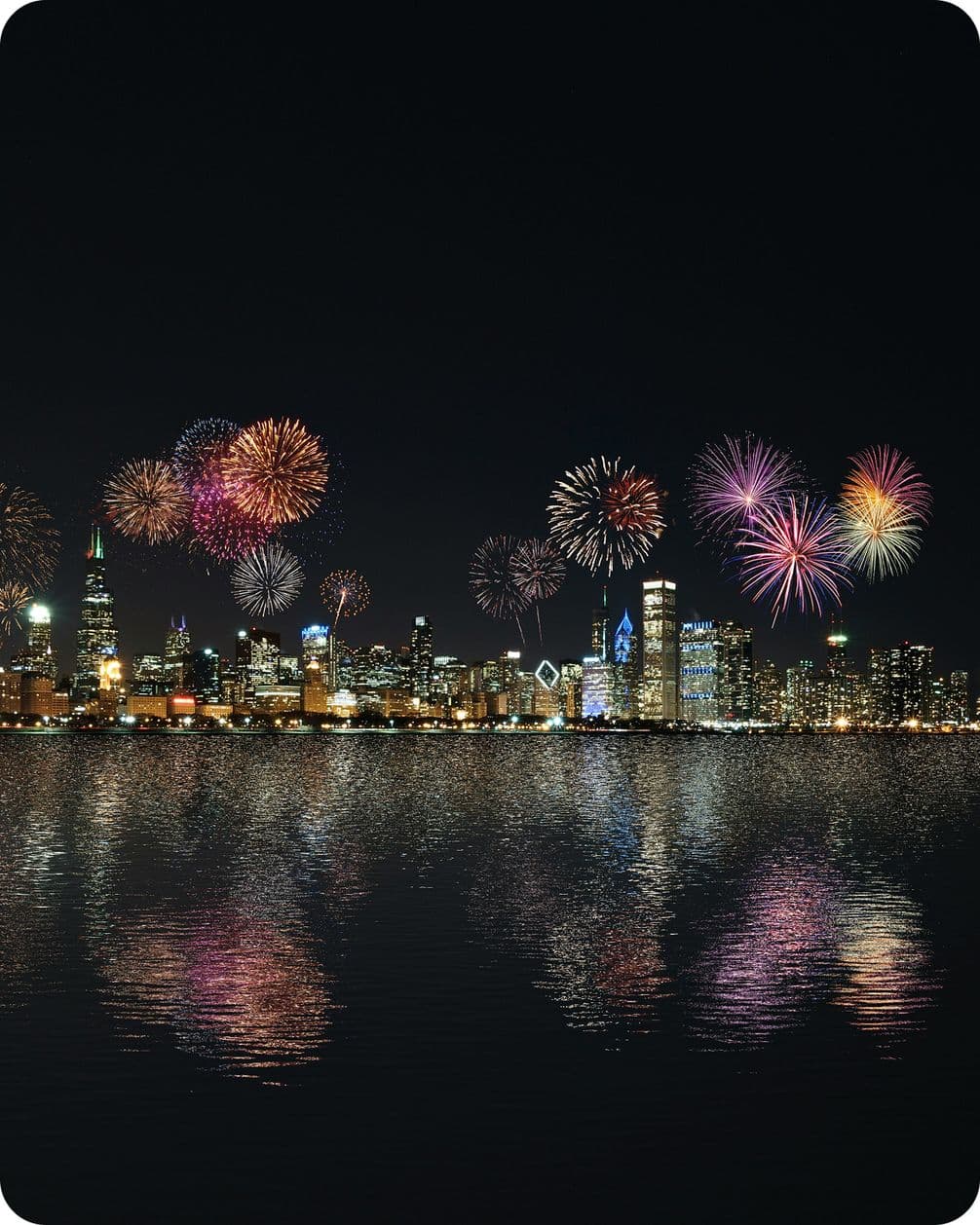Colorful fireworks illuminate a city skyline at night, reflecting vibrantly on the calm water below.