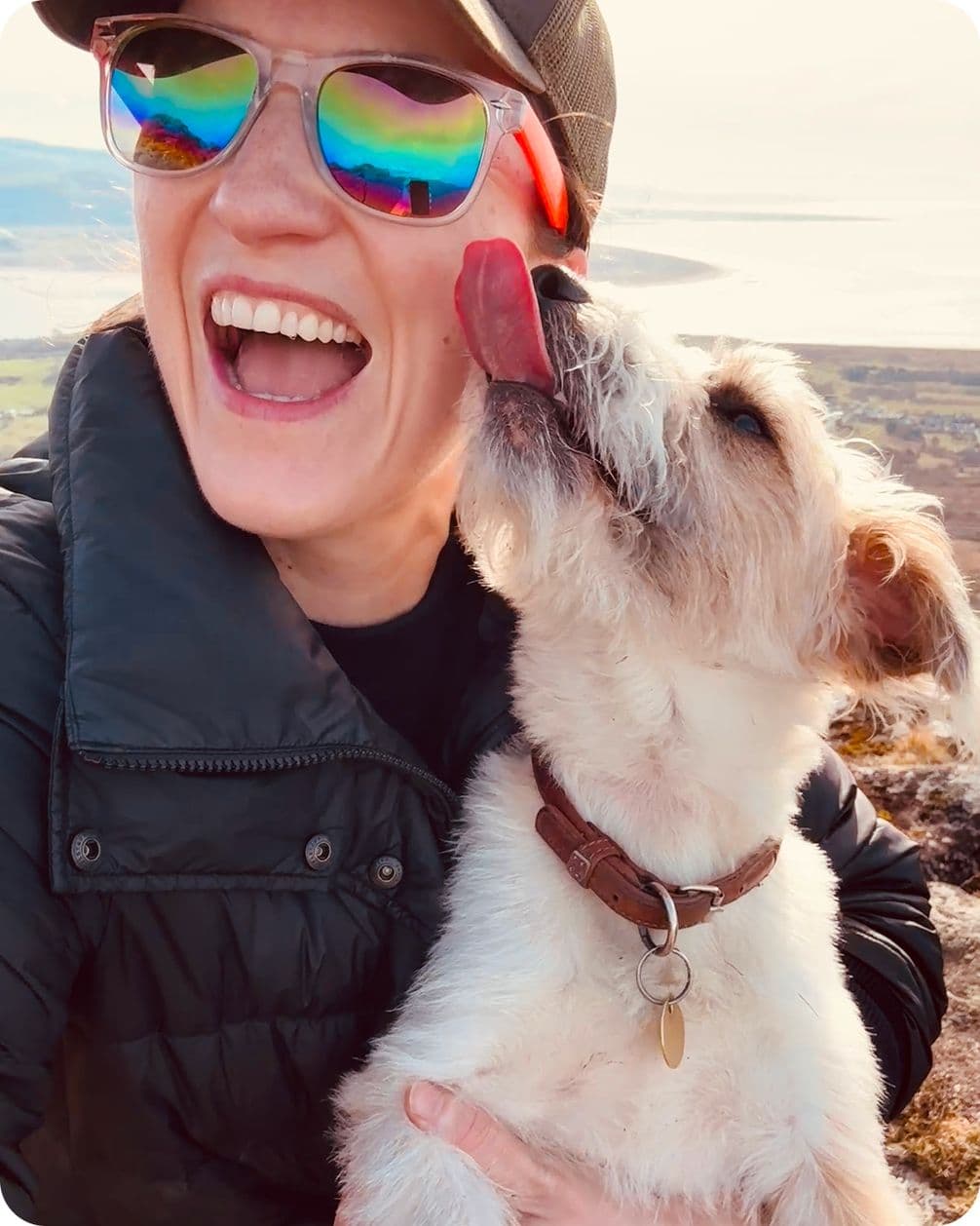 Smiling person in cap and reflective rainbow sunglasses laughs as a small white-and-brown dog licks their cheek outdoors.