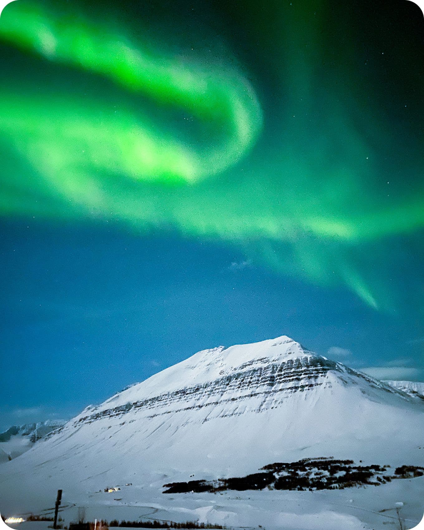 Northern lights swirl above a snow-covered mountain under a starry night sky, creating a vibrant green display.