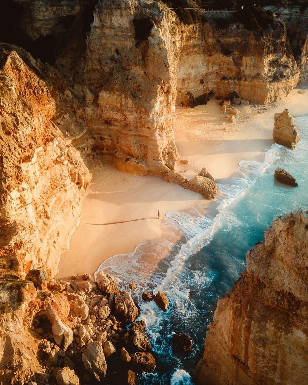 Aerial view of a secluded beach surrounded by tall cliffs, with waves gently lapping the sandy shore and a person walking along the water's edge.