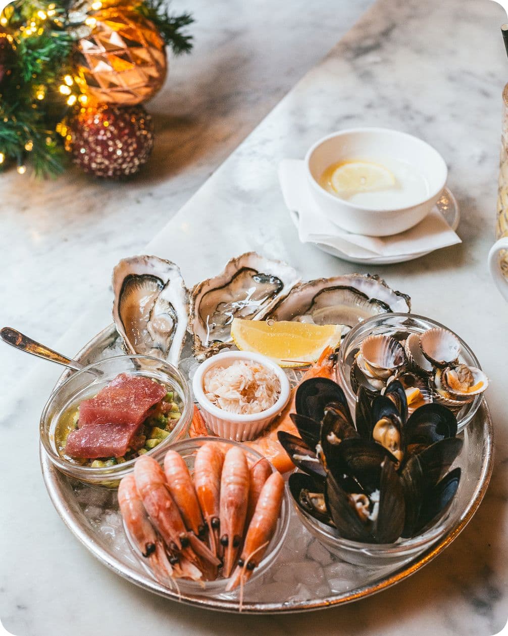 A seafood platter with oysters, shrimp, mussels, tuna, and clams on ice, garnished with lemon, served on a marble table.