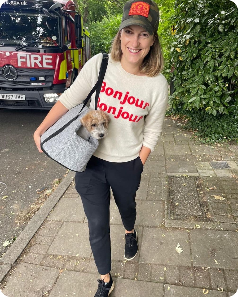 Smiling woman in 'Bonjour Bonjour' sweater and cap walks on sidewalk carrying small dog in gray carrier bag, fire engine parked behind.