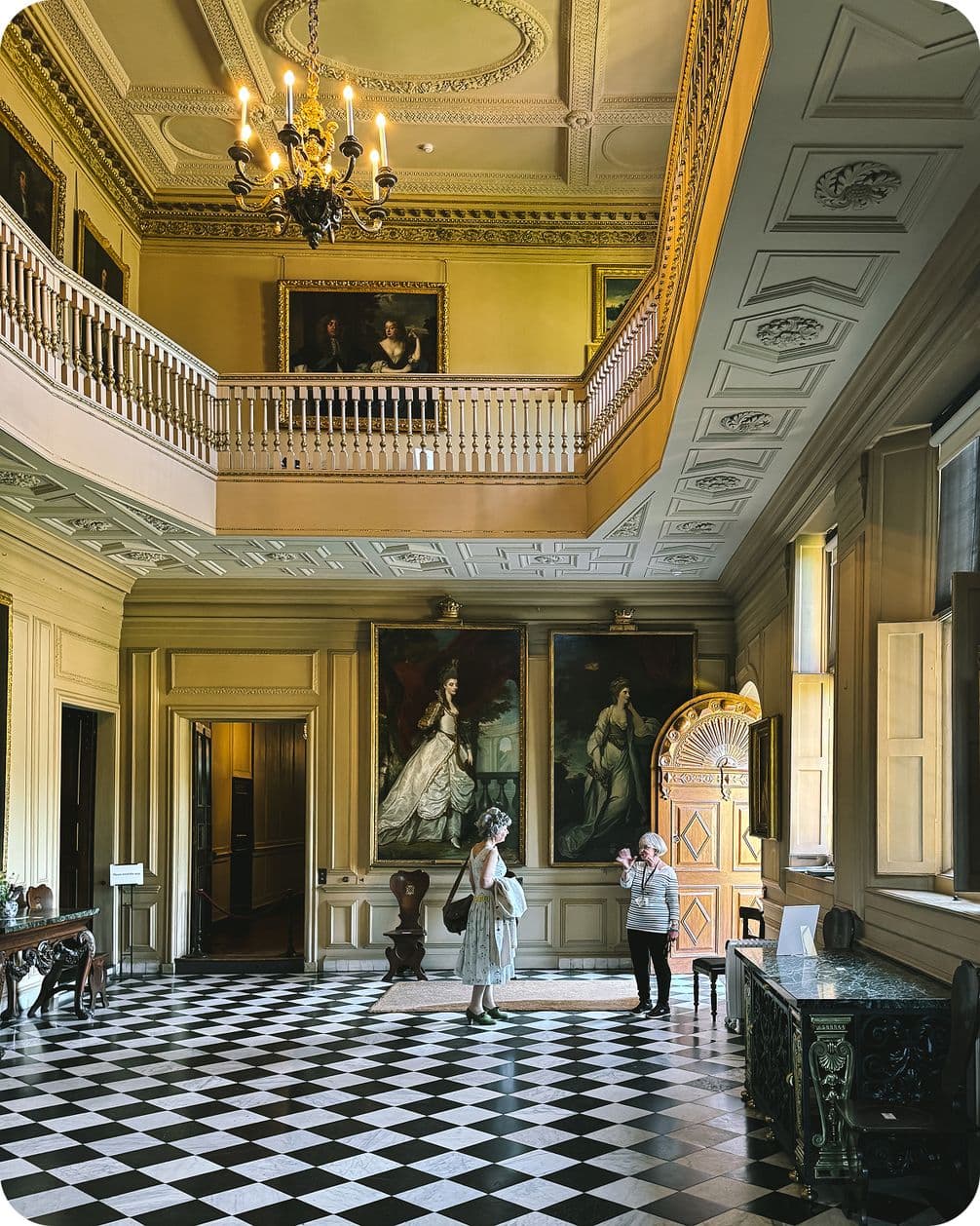 Elegant hall with checkered floor, ornate ceiling, chandelier, and large paintings. Two people stand near a window, illuminated by natural light.