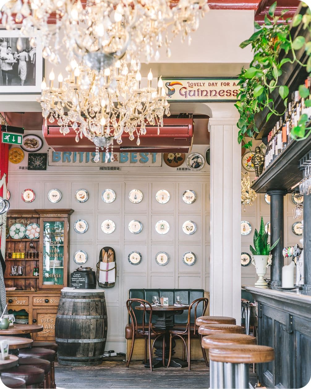 Cozy pub interior with chandeliers, vintage decor, wall plates, and a small table. A "Lovely Day for a Guinness" sign is displayed above.