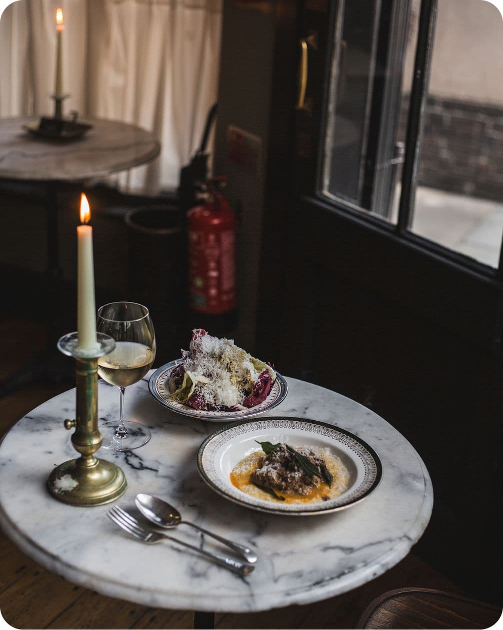 A marble table with a lit candle, wine glass, salad, and a main dish near a window in a dimly lit restaurant.