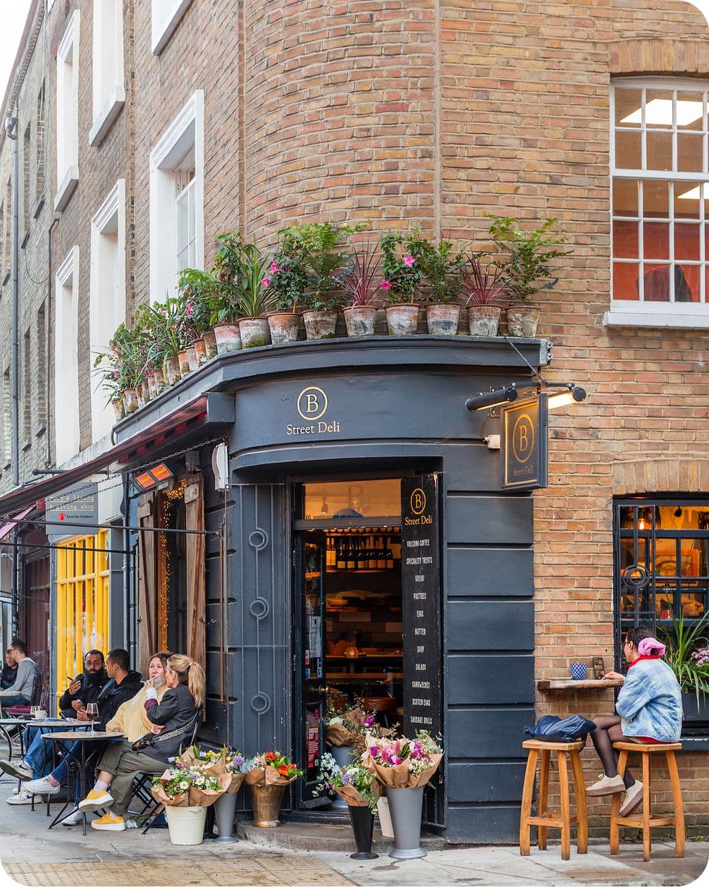 Street Deli on a corner with brick exterior, outdoor seating, potted plants above, and flower display. People seated, enjoying the atmosphere.