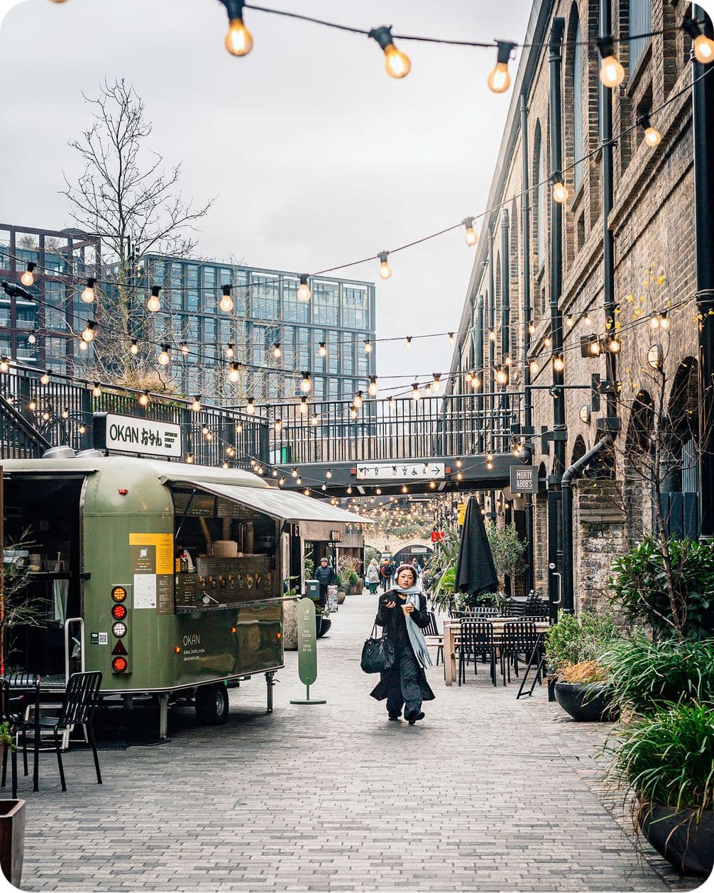 A woman walks through a lively outdoor market with string lights, food truck, and tables, set against urban buildings on a cloudy day.