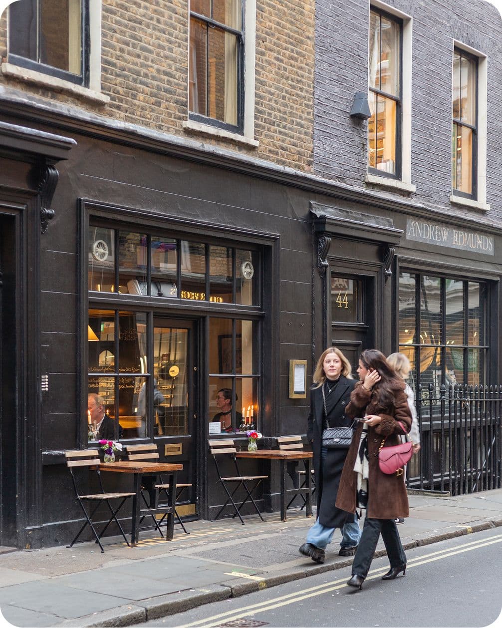 Two people walk past a cozy cafe with outdoor seating on a city street. The building has dark brick and large windows.