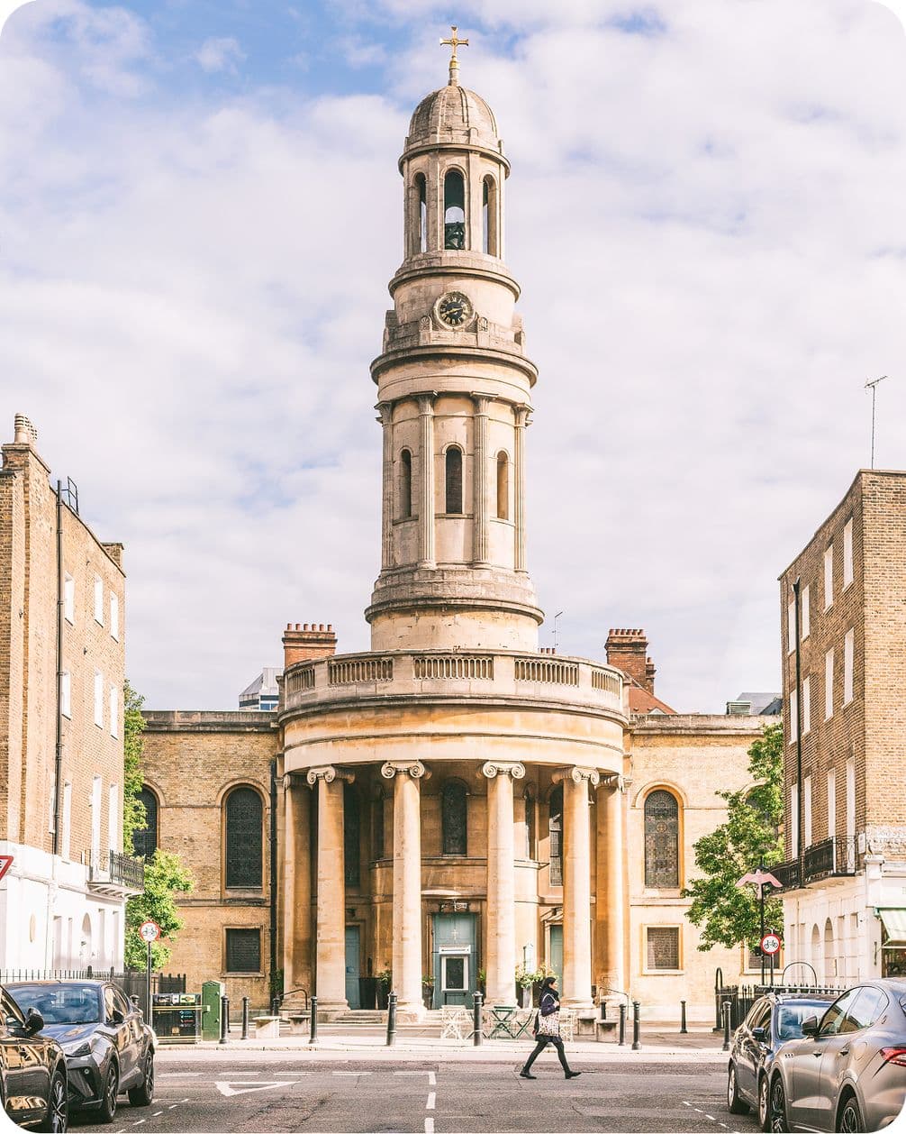 A person crosses the street in front of a historic church with a tall clock tower and columns, flanked by brick buildings under a cloudy sky.
