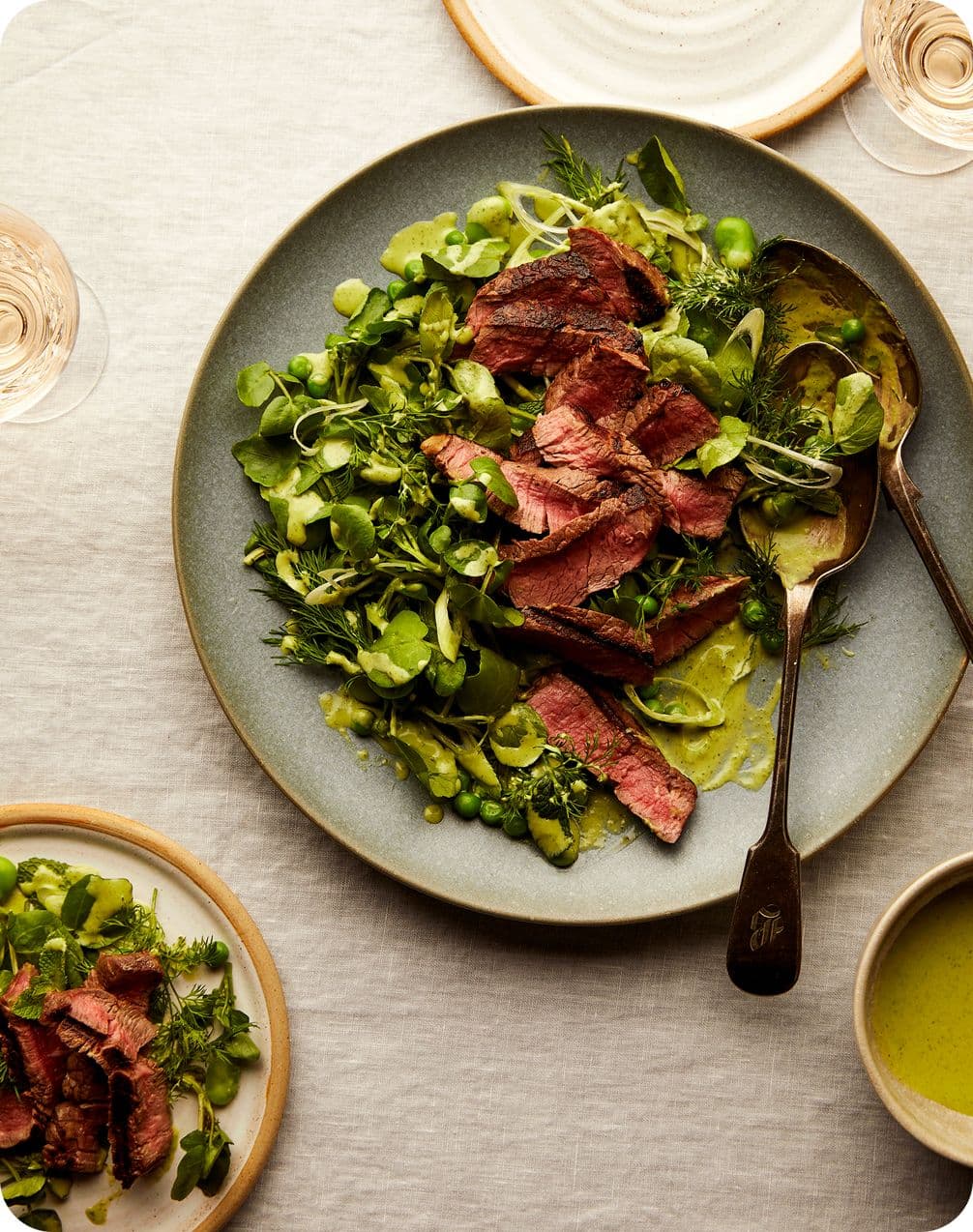 Top-down view of a plate of sliced medium-rare steak on a green pea and herb salad with a spoon, small bowls and wine glasses.