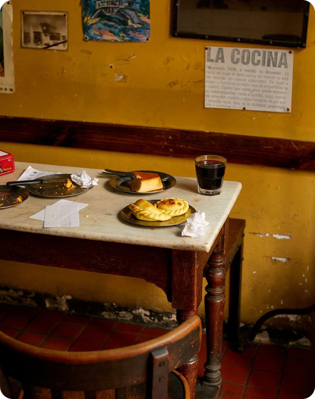 Marble-top cafe table with empanadas, a slice of cake, a glass of dark drink and plates against a worn yellow wall with a "La Cocina" sign.