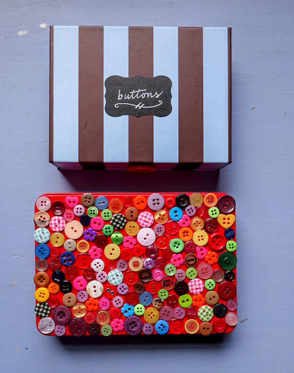 Striped box labeled 'buttons' above a tin lid densely covered with colorful buttons on a gray-blue background.