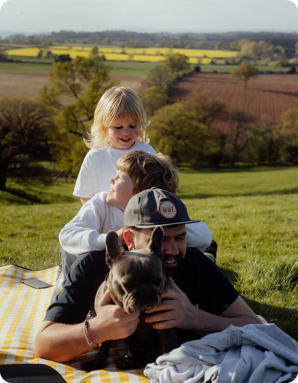 A man with a cap holds a dog while two children playfully sit on his back. They're on a picnic blanket in a scenic countryside field.