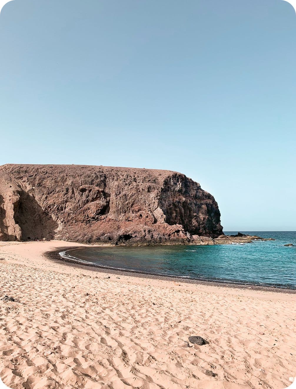 A serene beach with golden sand, turquoise water, and a rocky cliff under a clear blue sky.