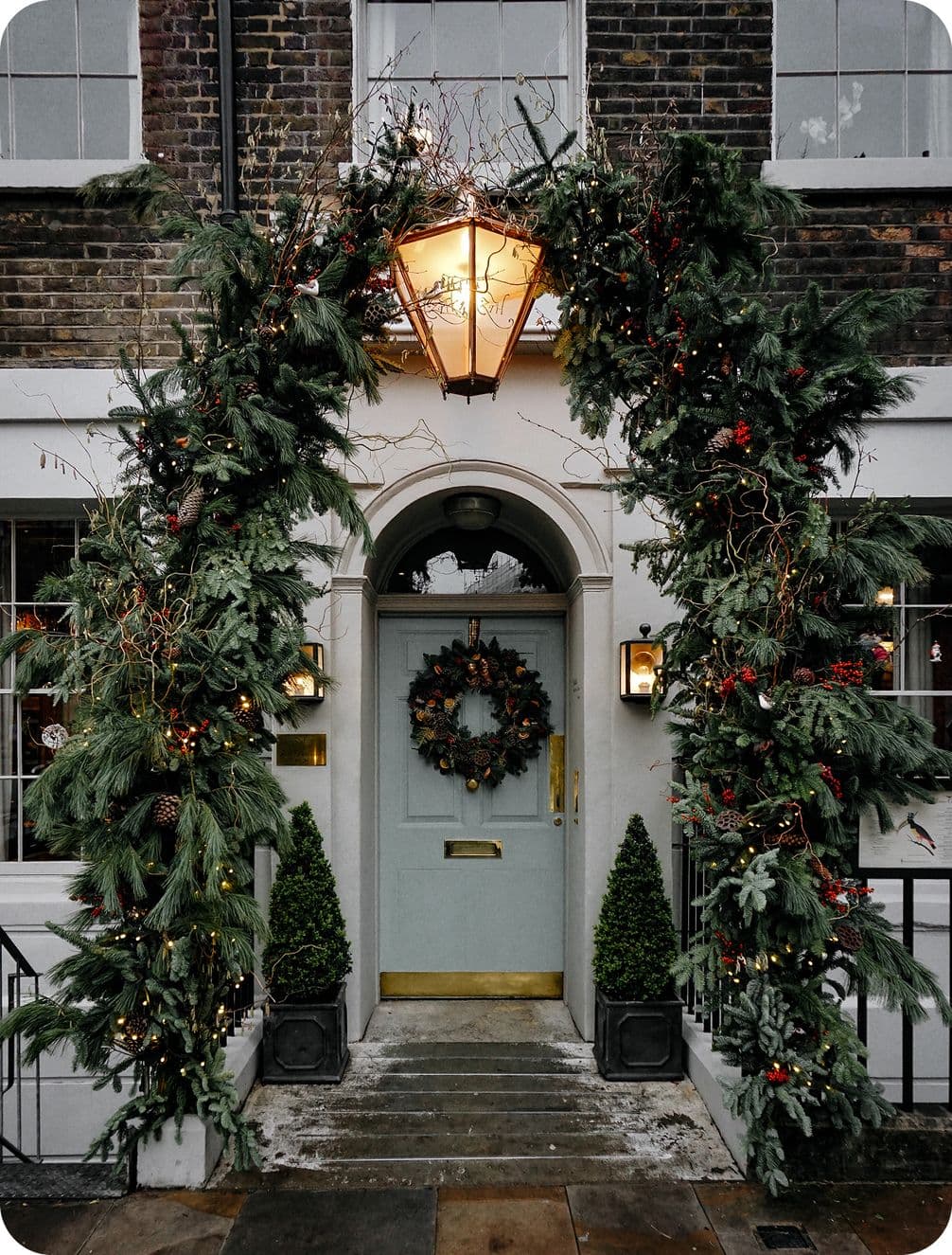 Festive doorway with pine garland and lights arching over it, a wreath on the door, and two small potted trees on either side.