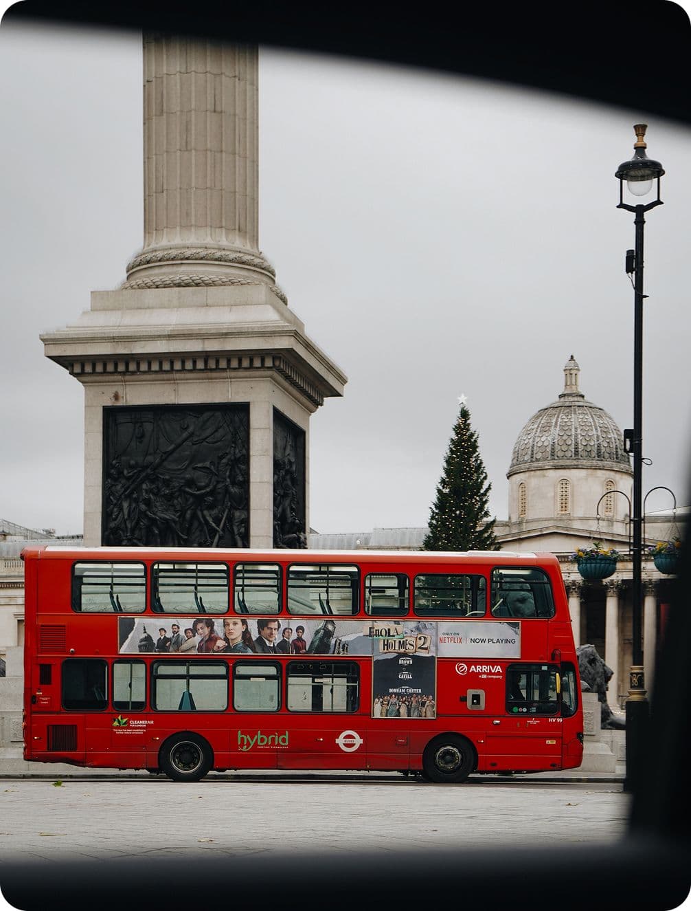 A red double-decker bus passes by a tall column and a domed building in a city square, with overcast skies above.