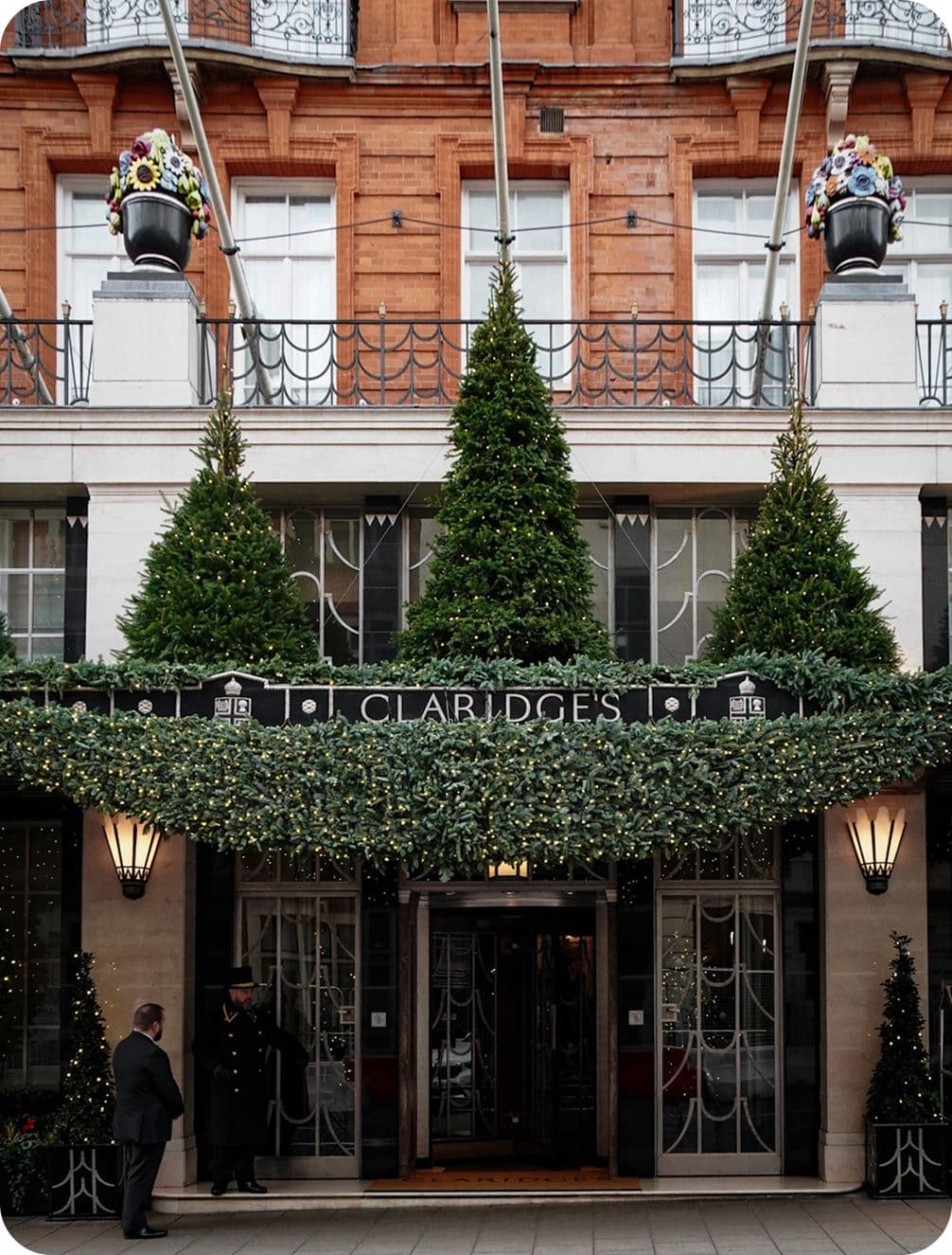 Entrance of a luxury hotel with festive decorations, including greenery, lights, and potted trees. Two people stand near the doorway.