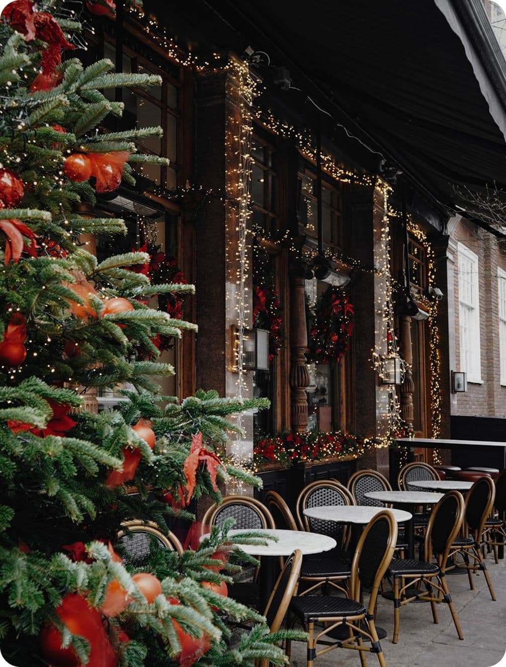 Festive outdoor café with decorated Christmas tree, string lights, and empty tables set against a brick building backdrop.
