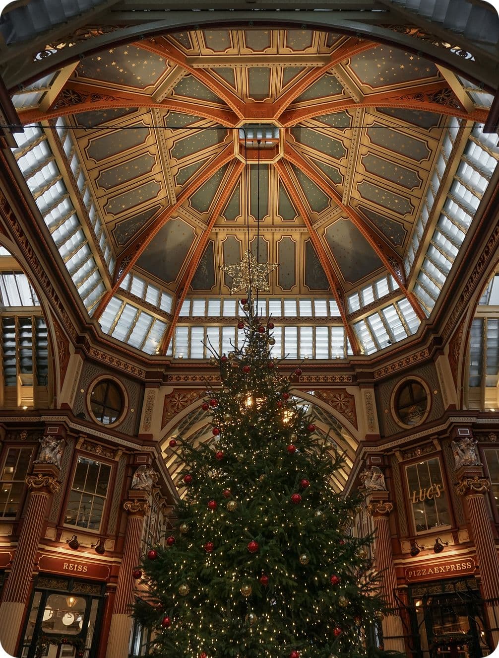 A tall Christmas tree with lights stands under an ornate, domed ceiling in a decorated indoor shopping area.