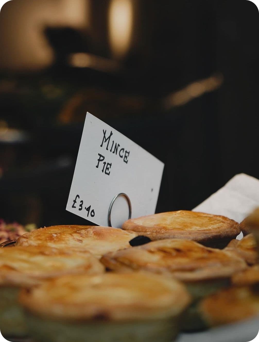 Close-up of golden-brown mince pies on display with a sign reading "Mince Pie £3.40" in a bakery setting.