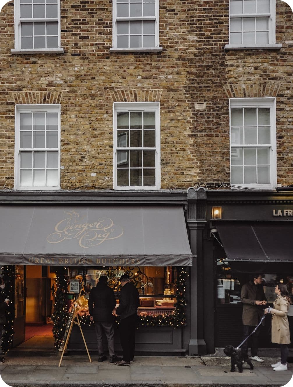 People standing outside a quaint butcher shop with festive decorations, next to a dark storefront. A person walks a dog on the sidewalk.