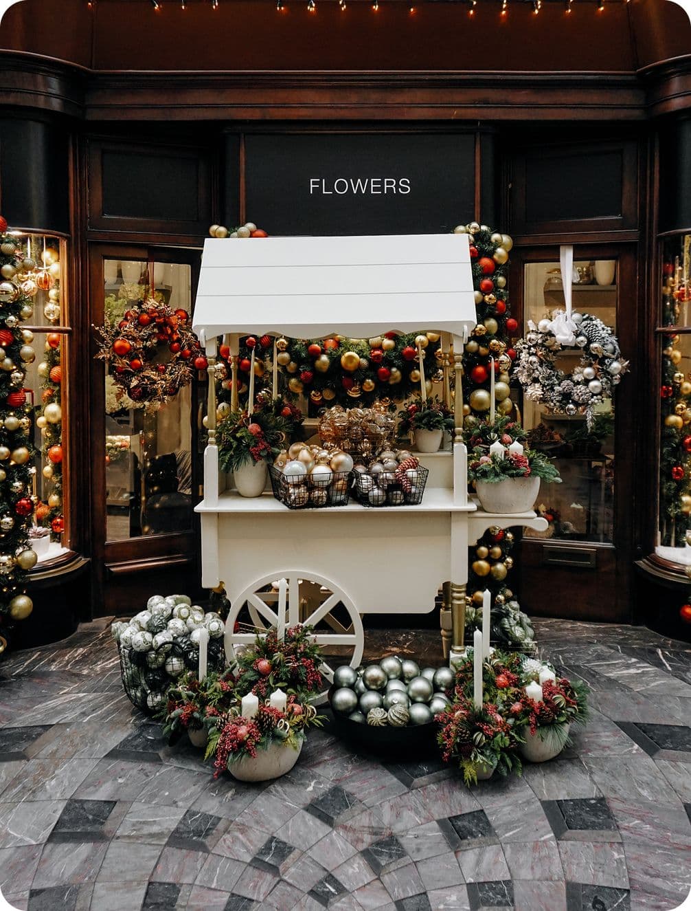 A festive flower cart adorned with Christmas ornaments and wreaths, surrounded by decorated windows and holiday greenery.