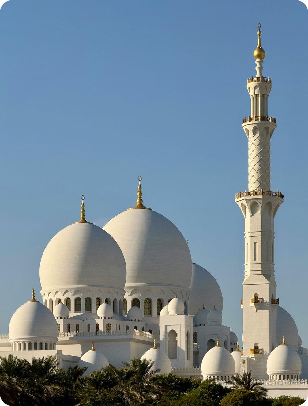 A grand mosque with large white domes and a tall minaret, set against a clear blue sky, surrounded by palm trees.