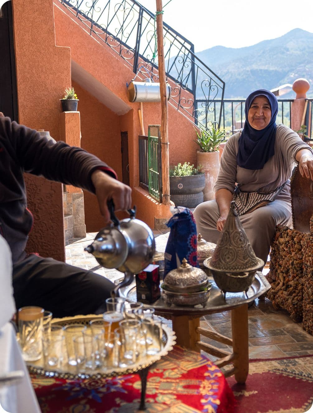 A person pours tea from a kettle at an outdoor table with ornate teapots and glasses. Another person sits smiling, with mountains in the background.