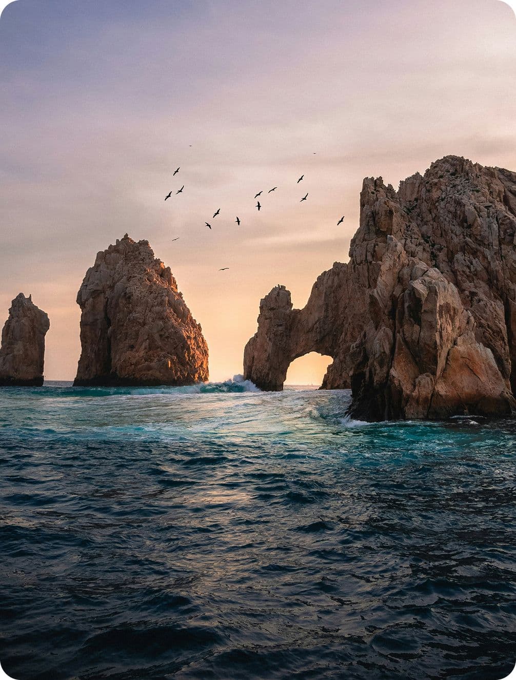 Rocky arch formations rise from the ocean at sunset, with birds flying overhead and waves crashing below.