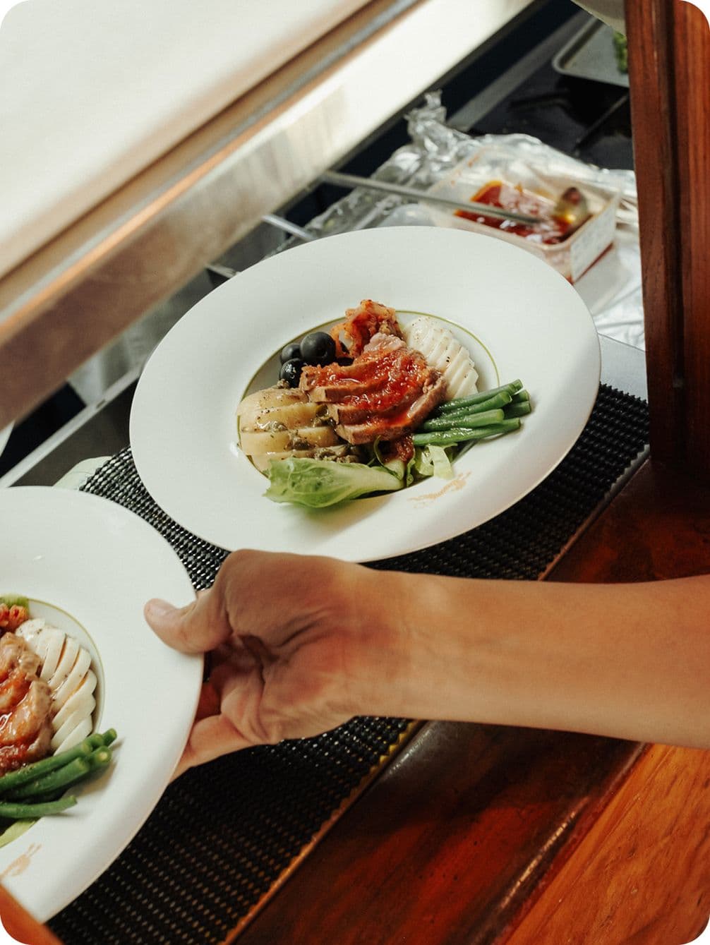 A hand serves a plated meal with sliced meat, green beans, olives, and lettuce on a white plate, set on a woven mat.