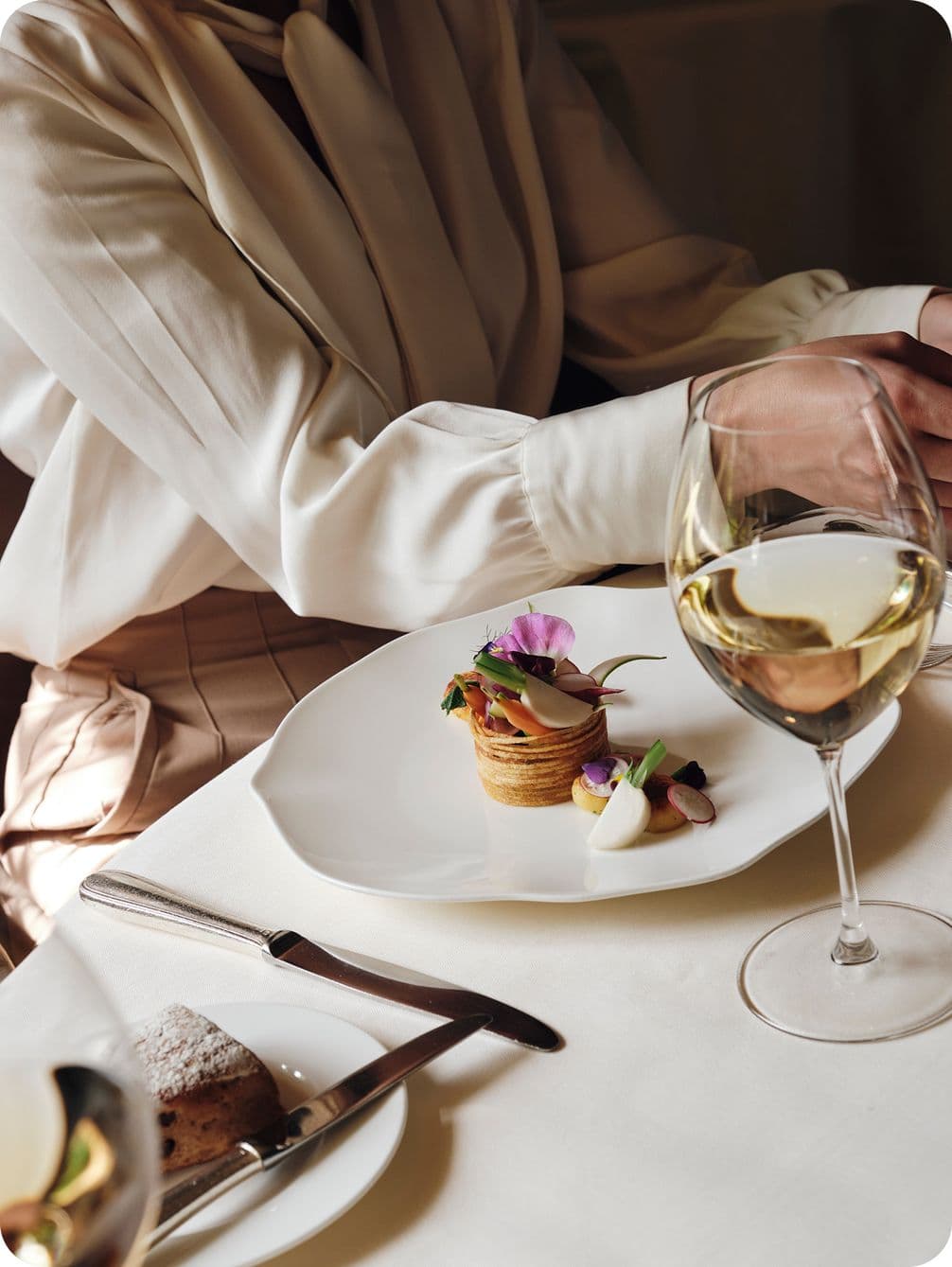 A person in a white blouse sits at a table with a gourmet dish and a glass of white wine, with cutlery and bread beside them.