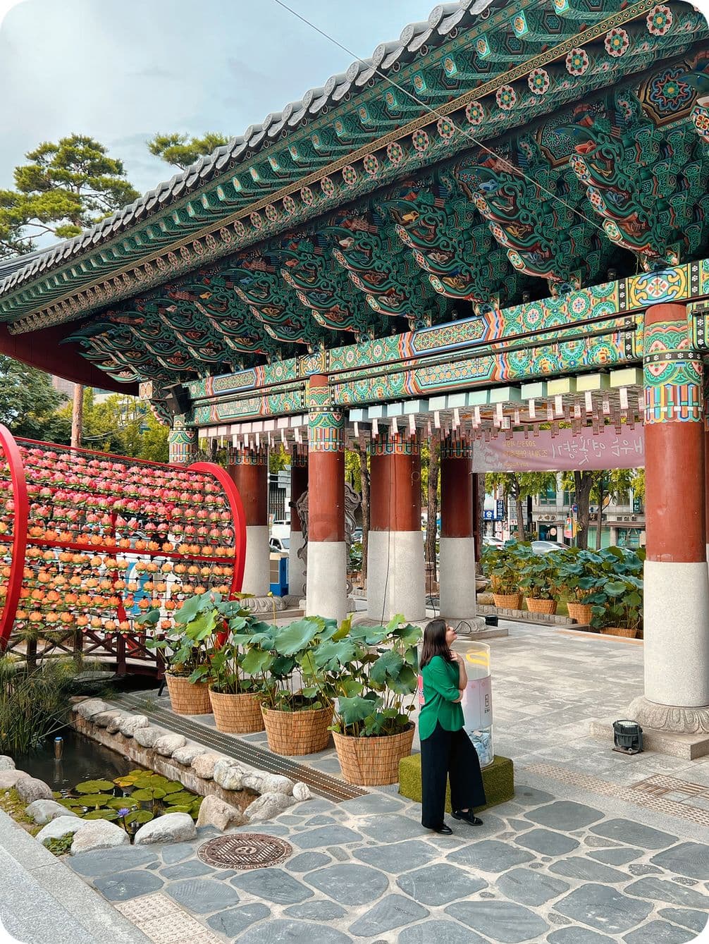 A woman stands near a traditional temple gate adorned with colorful lanterns and intricate designs, surrounded by potted plants and lotus flowers.