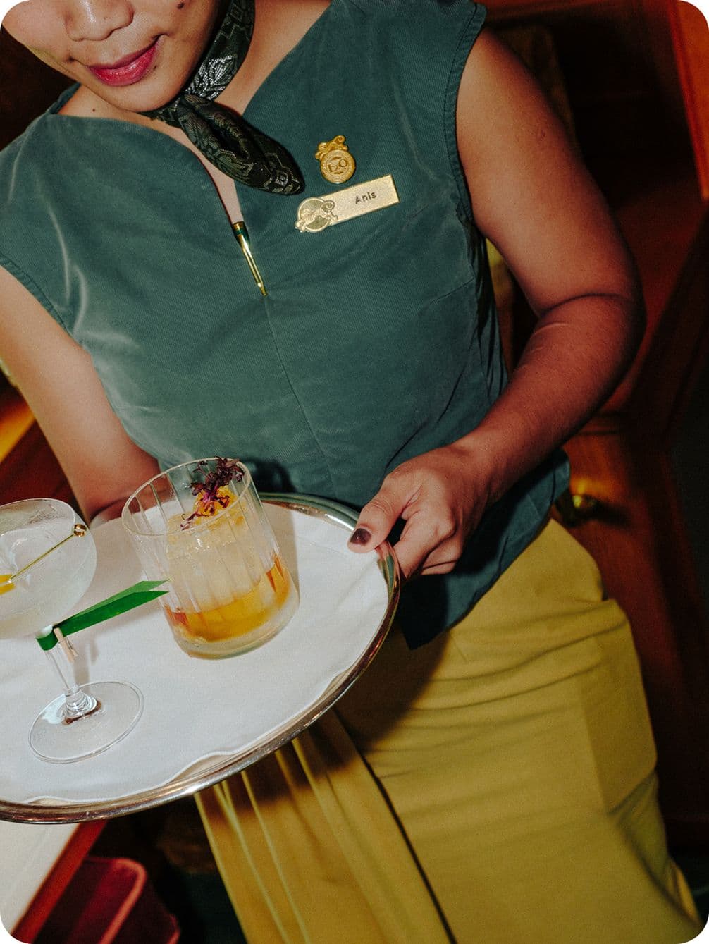 Person holding a tray with two cocktails, wearing a green top and yellow skirt, with a name tag and scarf.