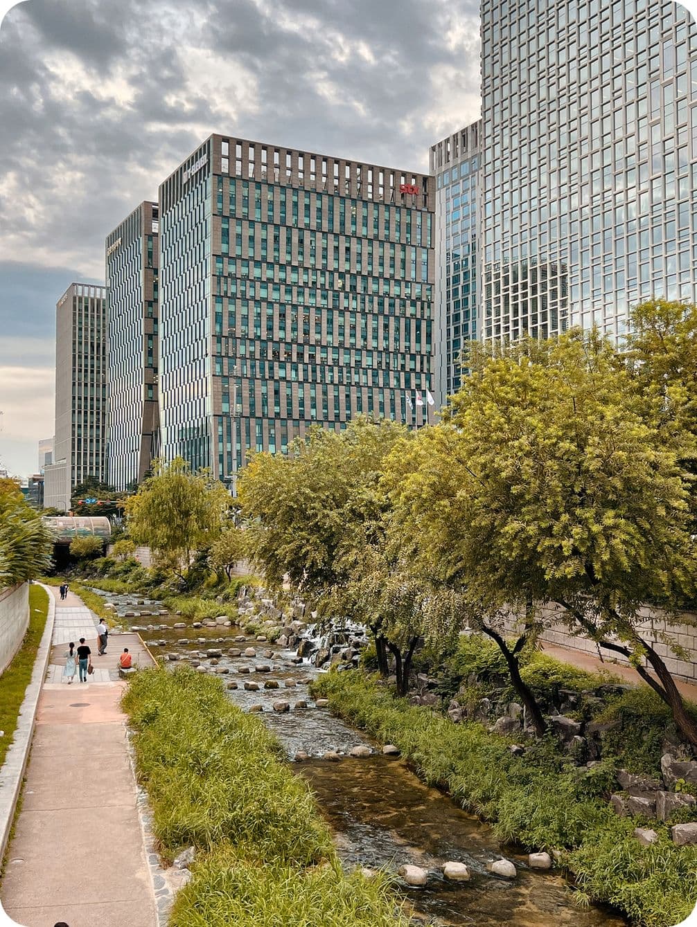 Modern buildings line a river with stepping stones, bordered by trees and a pathway where people walk, under a cloudy sky.