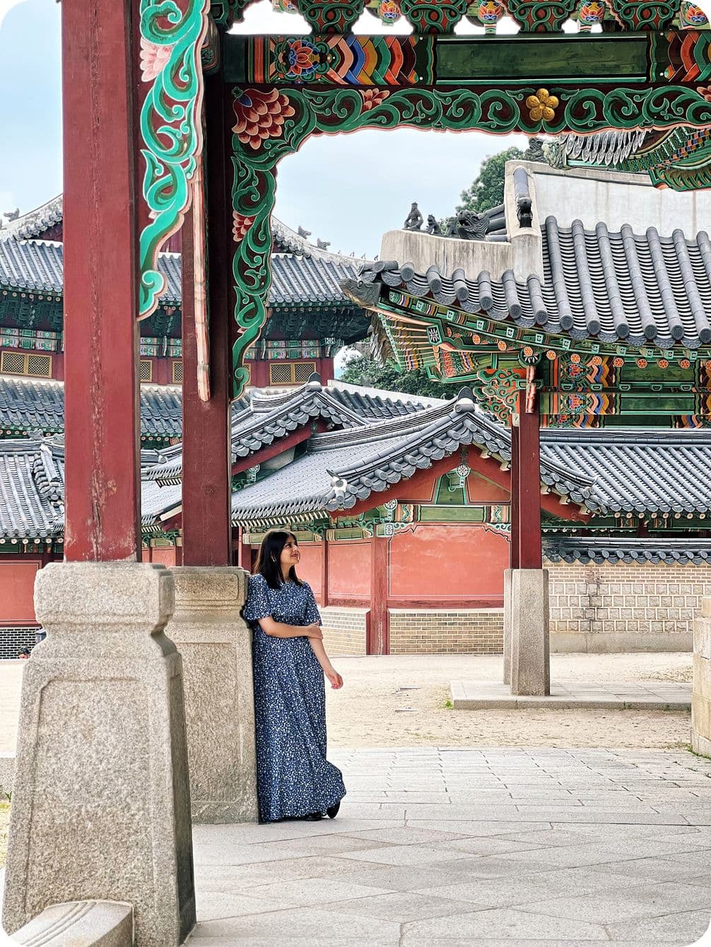Woman in blue dress stands under ornate, traditional Korean architecture with colorful wooden details and tiled roofs.