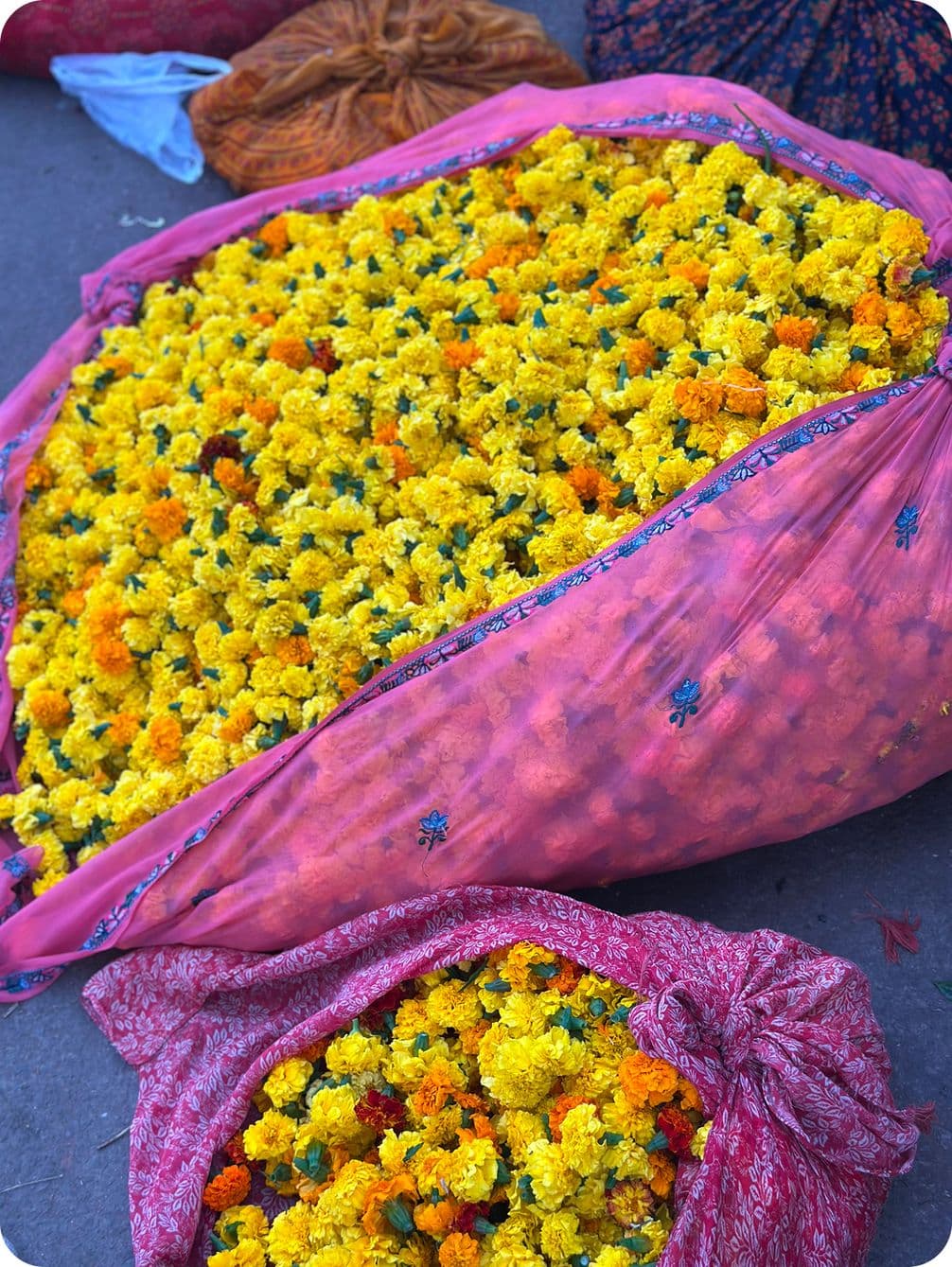 A large pink cloth bag filled with vibrant yellow and orange marigold flowers, with another smaller red cloth bag beside it.