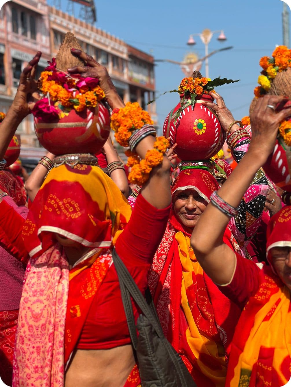 Women in vibrant red and yellow sarees carry decorated pots on their heads during a cultural procession, adorned with flowers and ornaments.