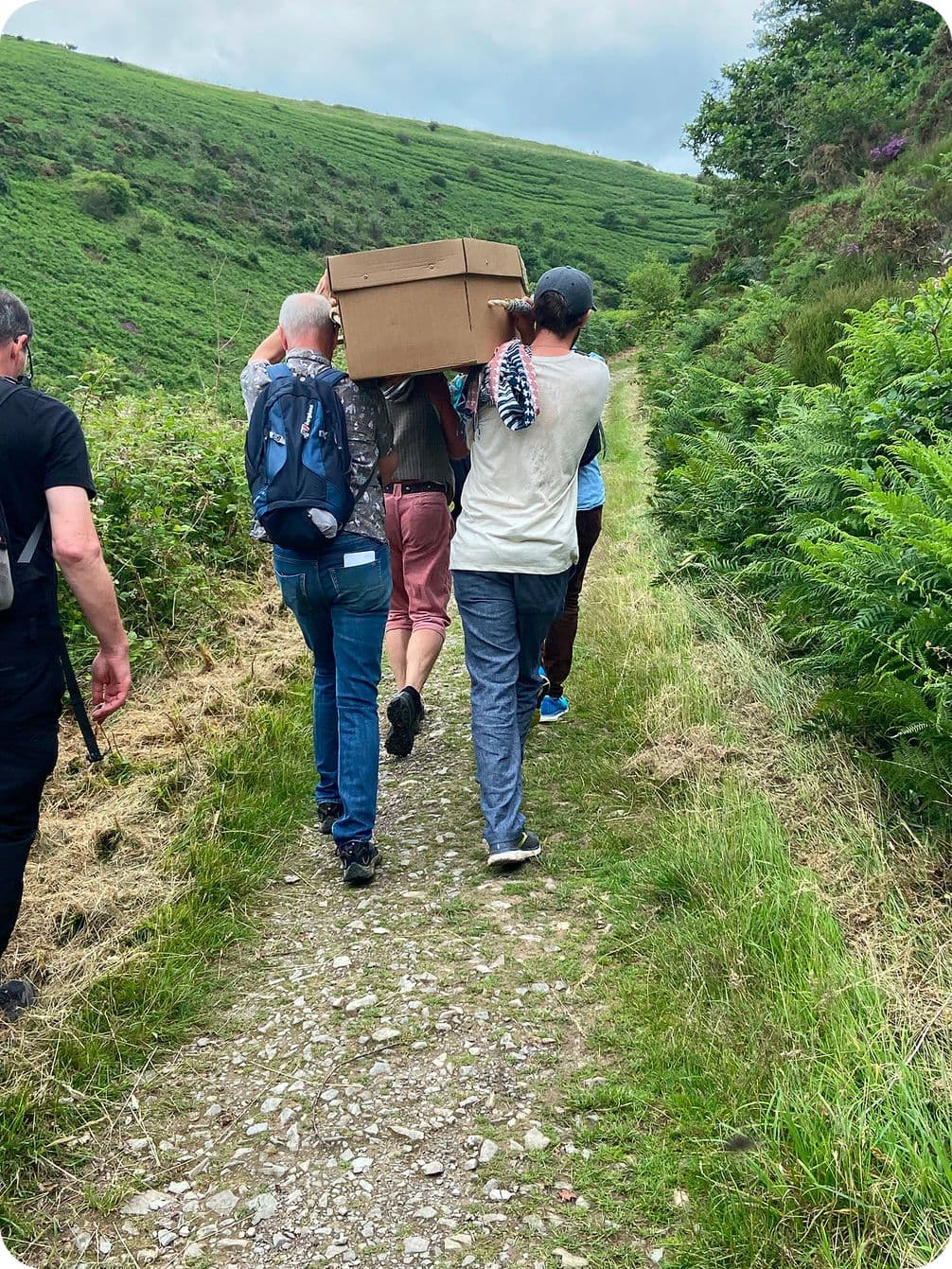 Four people walking a dirt path through green hills, two shouldering a large cardboard box.