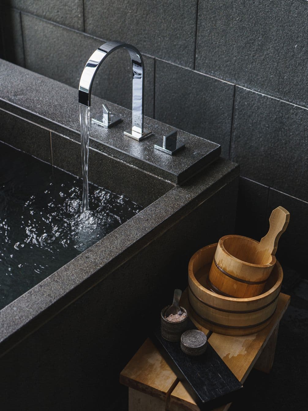 Modern stone bathtub with water flowing from a sleek faucet, wooden bucket and bowls on a small bench, set against dark tiled walls.