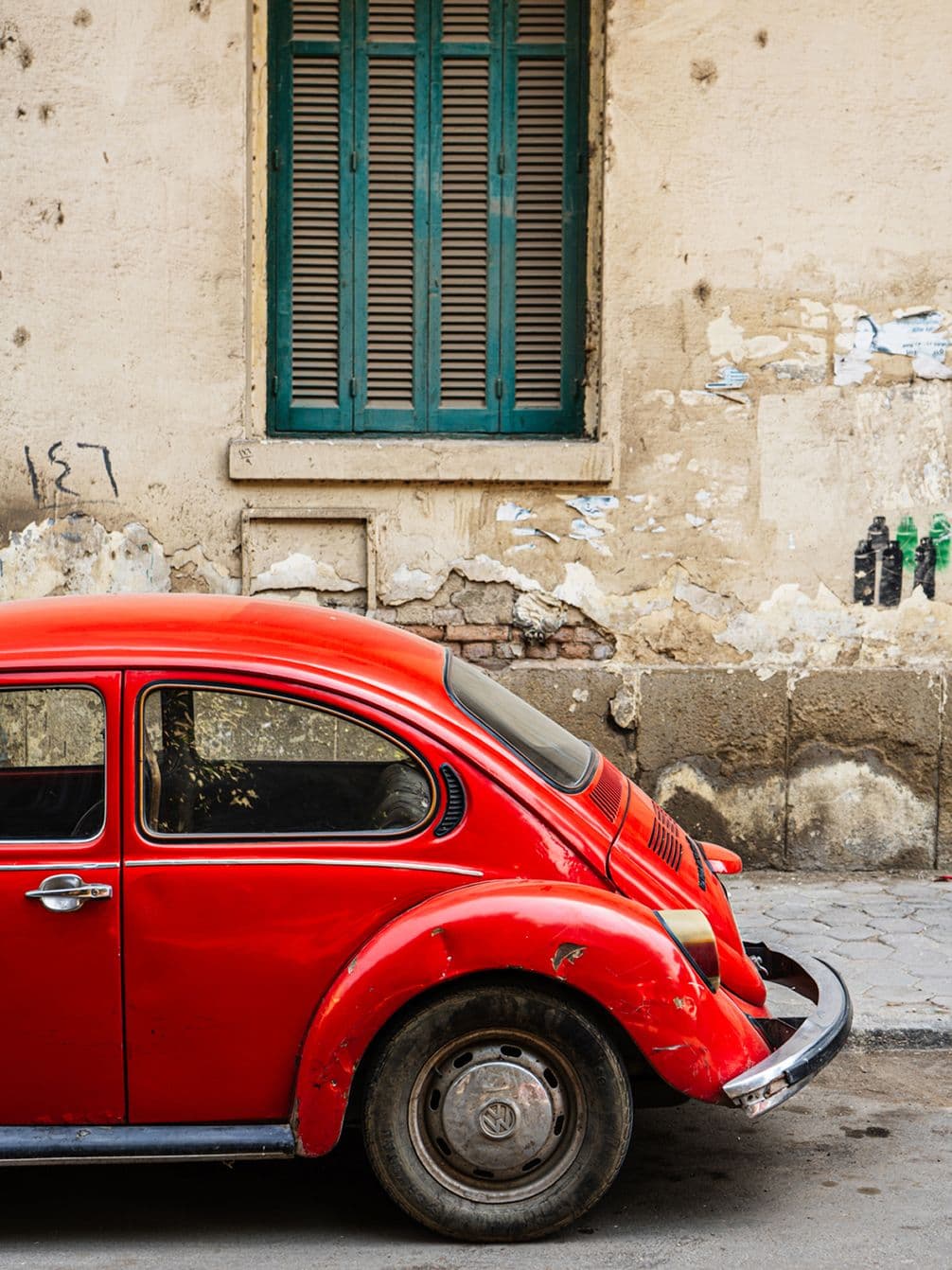 A red vintage car parked on a street in front of an old beige wall with a closed teal window. Peeling paint and a cracked sidewalk are visible.