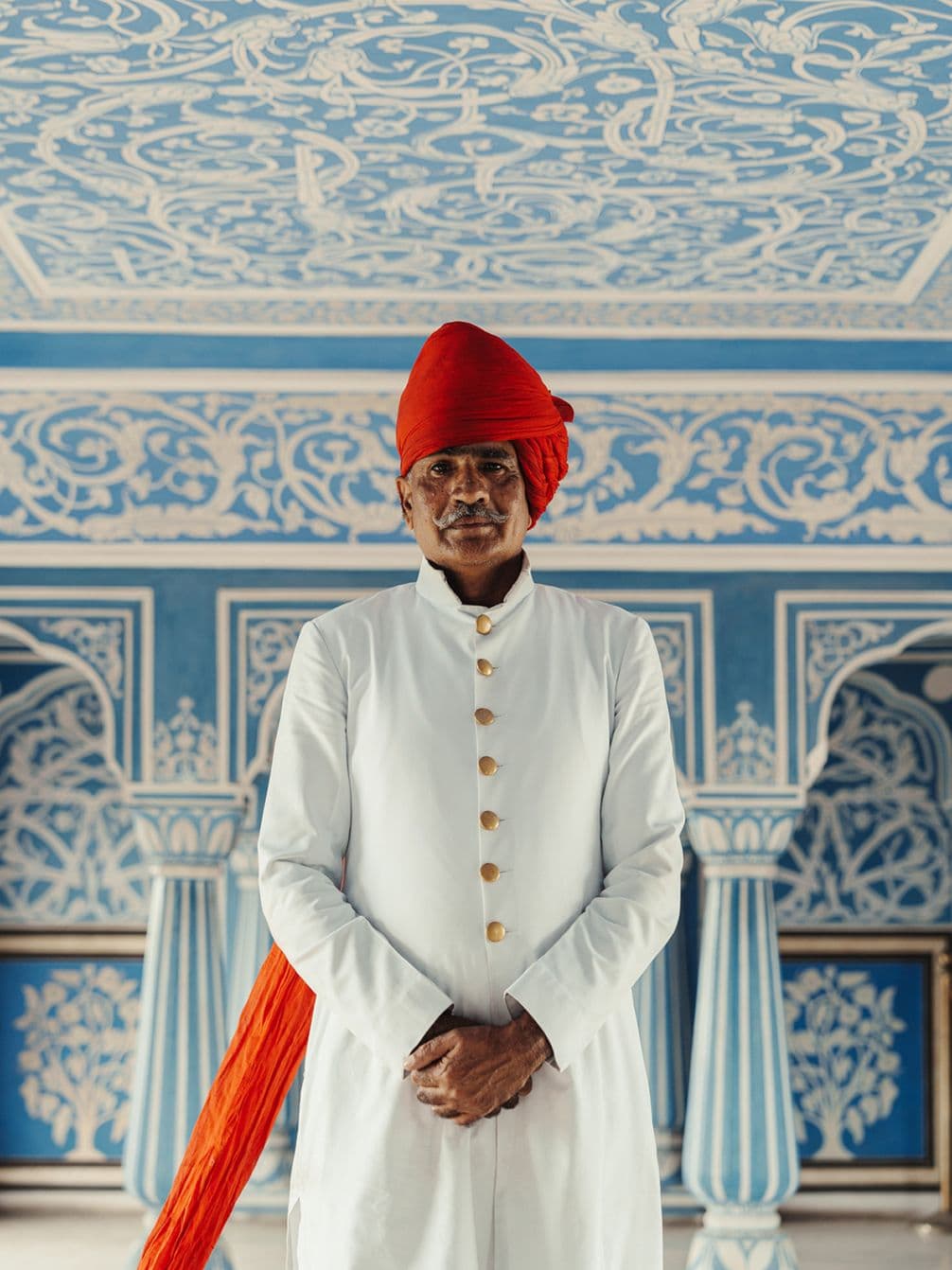 Man in white traditional attire with red turban stands in ornately decorated blue room with intricate patterns.