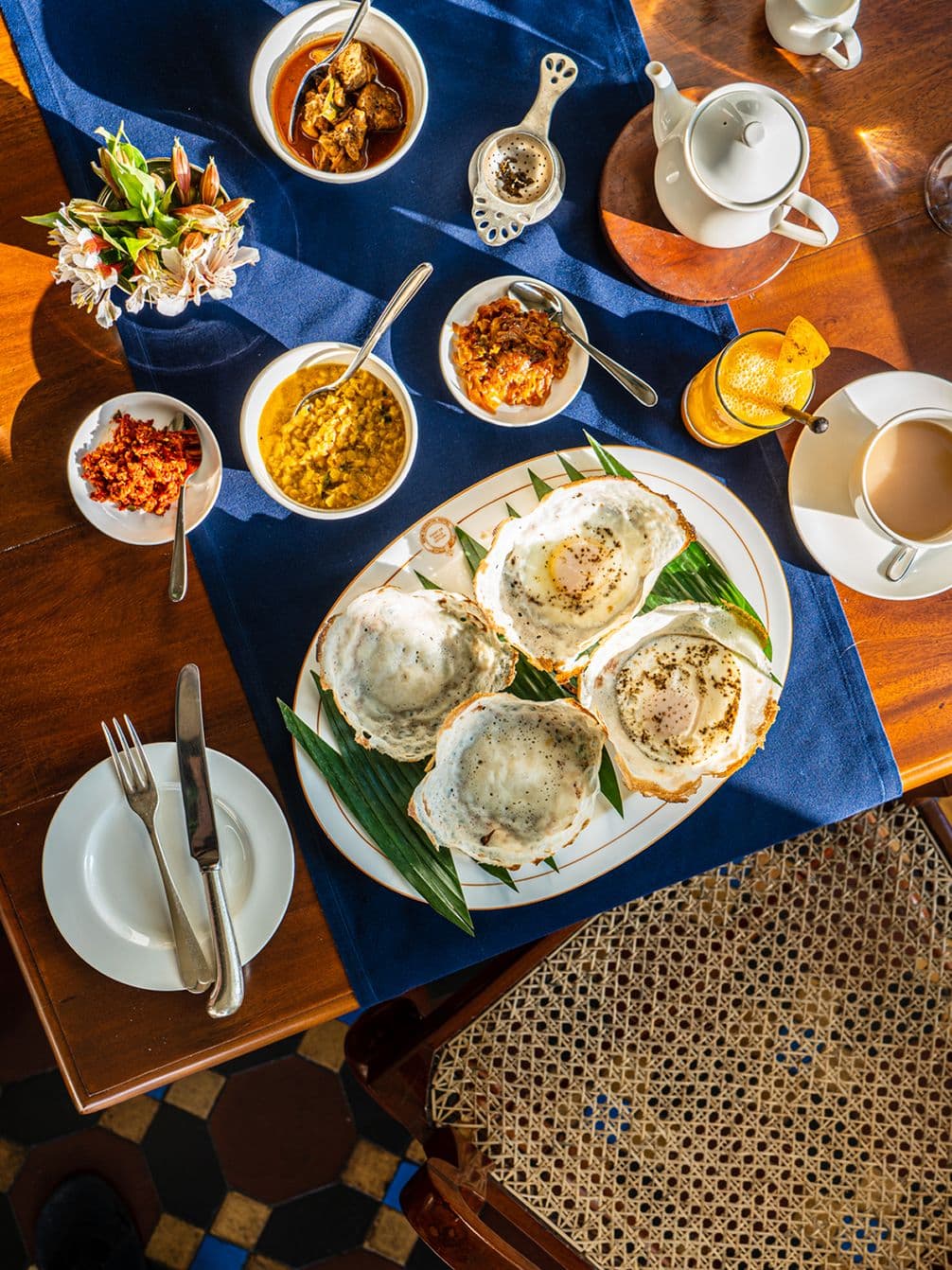 Aerial view of a wooden table with a breakfast spread including hoppers, curries, tea, and flowers on a blue tablecloth.