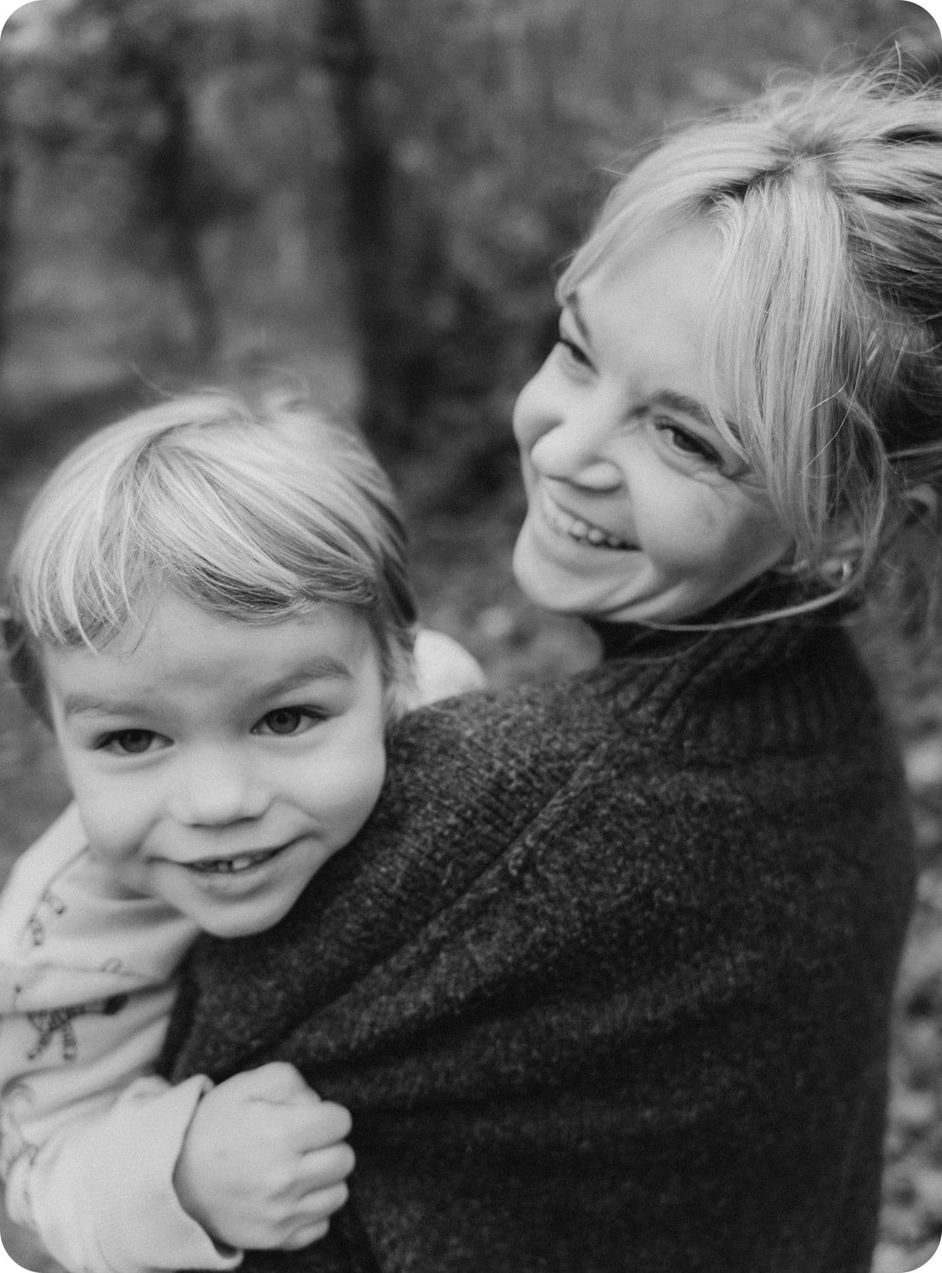 Black and white photo of a smiling woman holding a young child outdoors. Both are looking to the right, with trees blurred in the background.