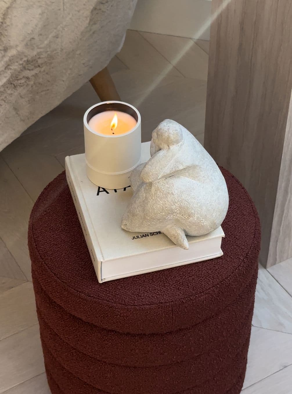 A lit candle in a white holder sits atop two books with a small stone sculpture of a contemplative figure beside them on a textured stool.