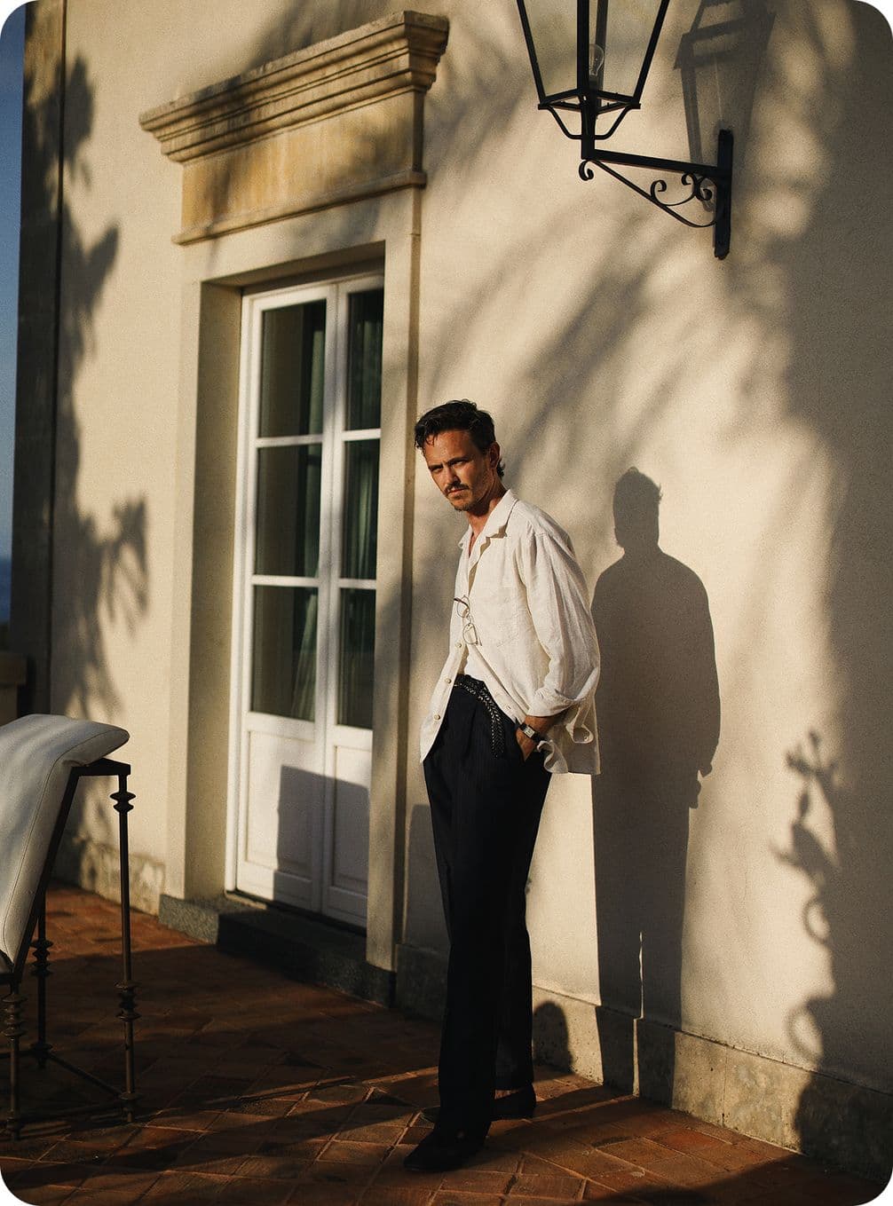 Man in a white shirt and dark pants stands on a sunlit patio, casting a shadow on a wall with a door and lantern.